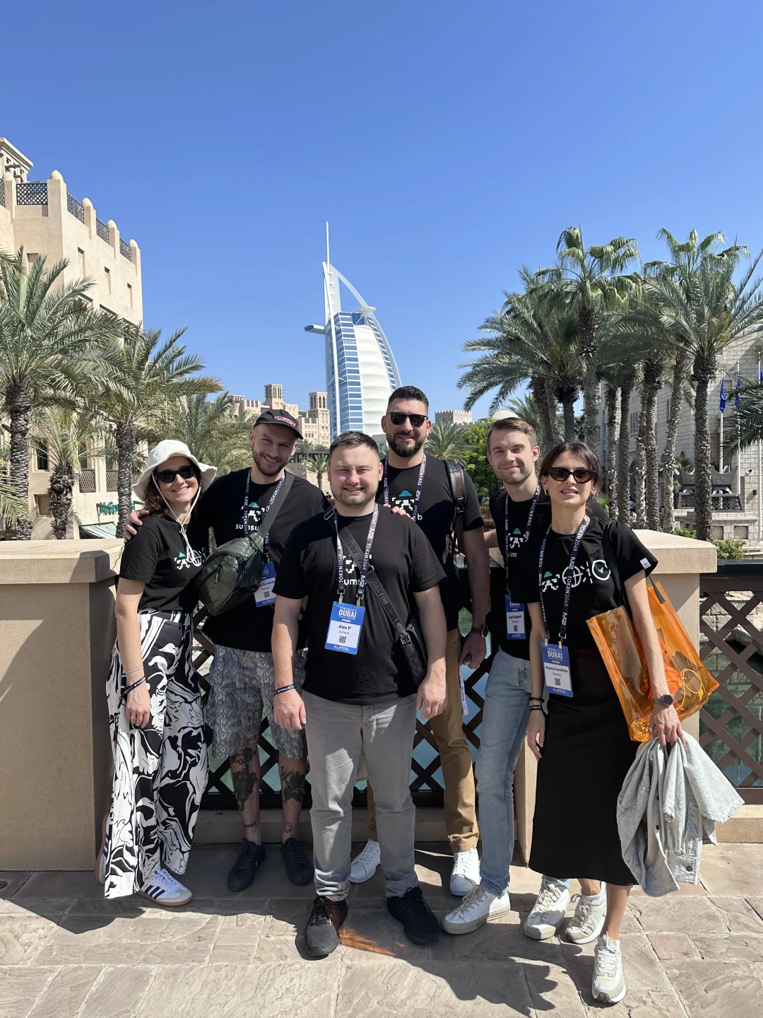 Six Sumsub team members with lanyards posing outdoors among palm trees, a sail-shaped tower behind them