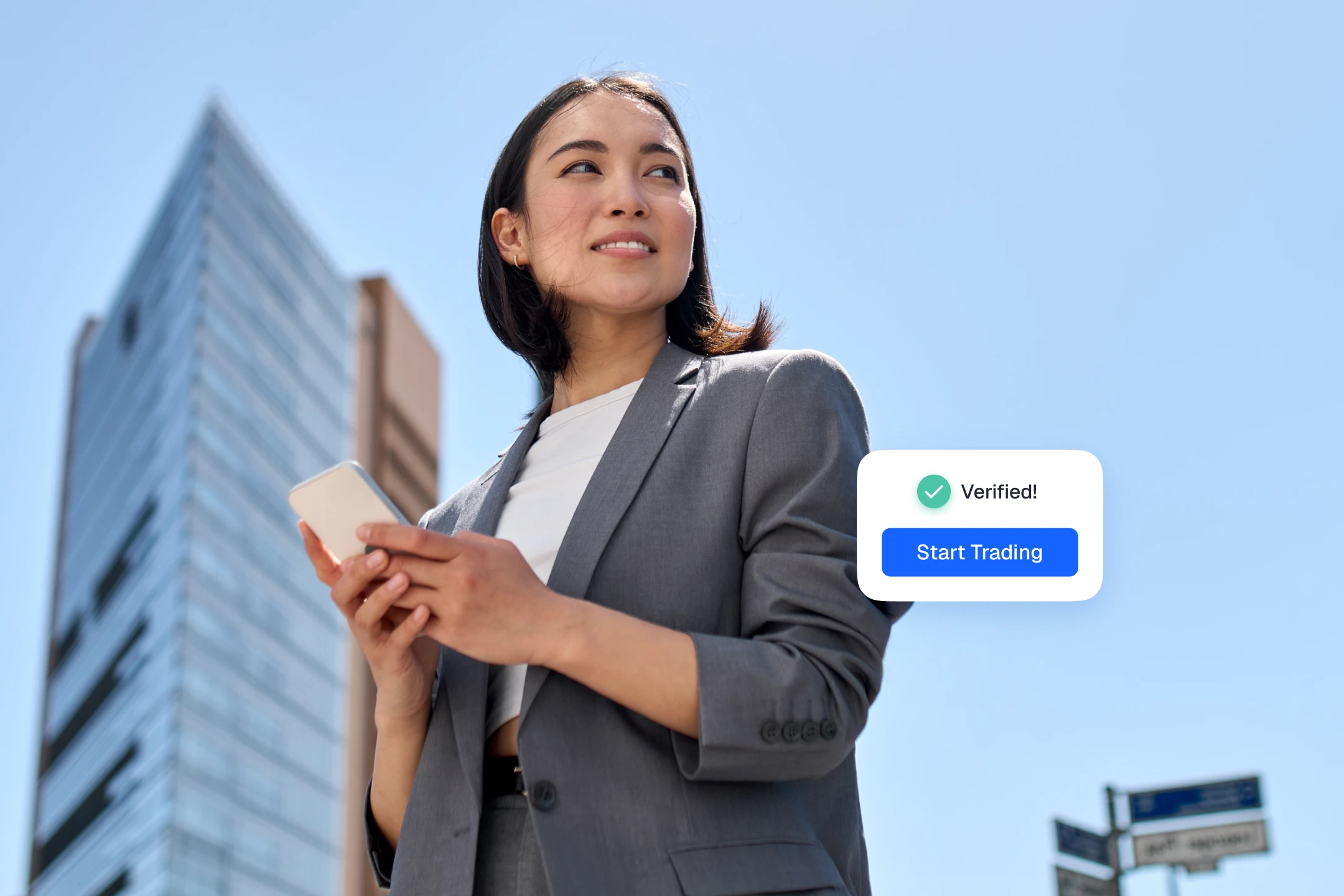Woman in a gray blazer holding a phone beside a "Verified! Start Trading" notification in front of a glass tower